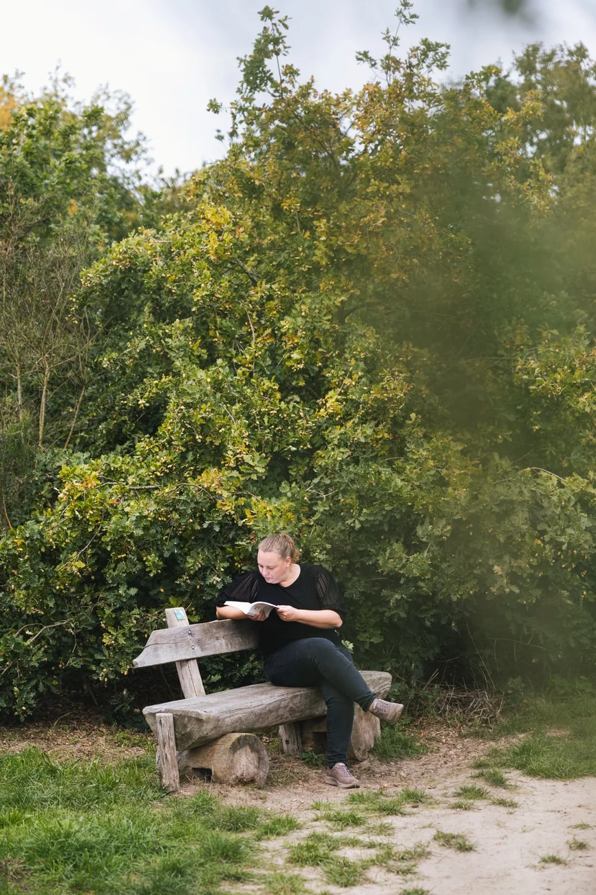 Lindsy leest een boek op een houten bank tussen de bomen.